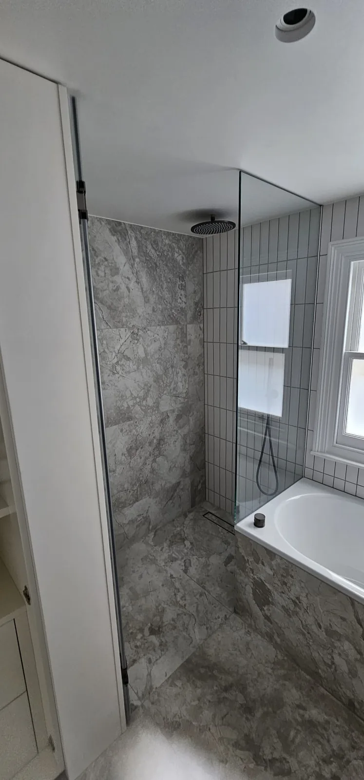Walk-in shower beside the bath: Large-format tiles, a frameless glass screen, a linear drain and a rain head.
